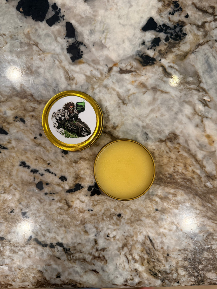 Open gold tin of Bombshell Suds Co. Sacred Ground beard balm showing the golden balm, next to its closed lid with a mad-scientist character graphic, on a granite countertop