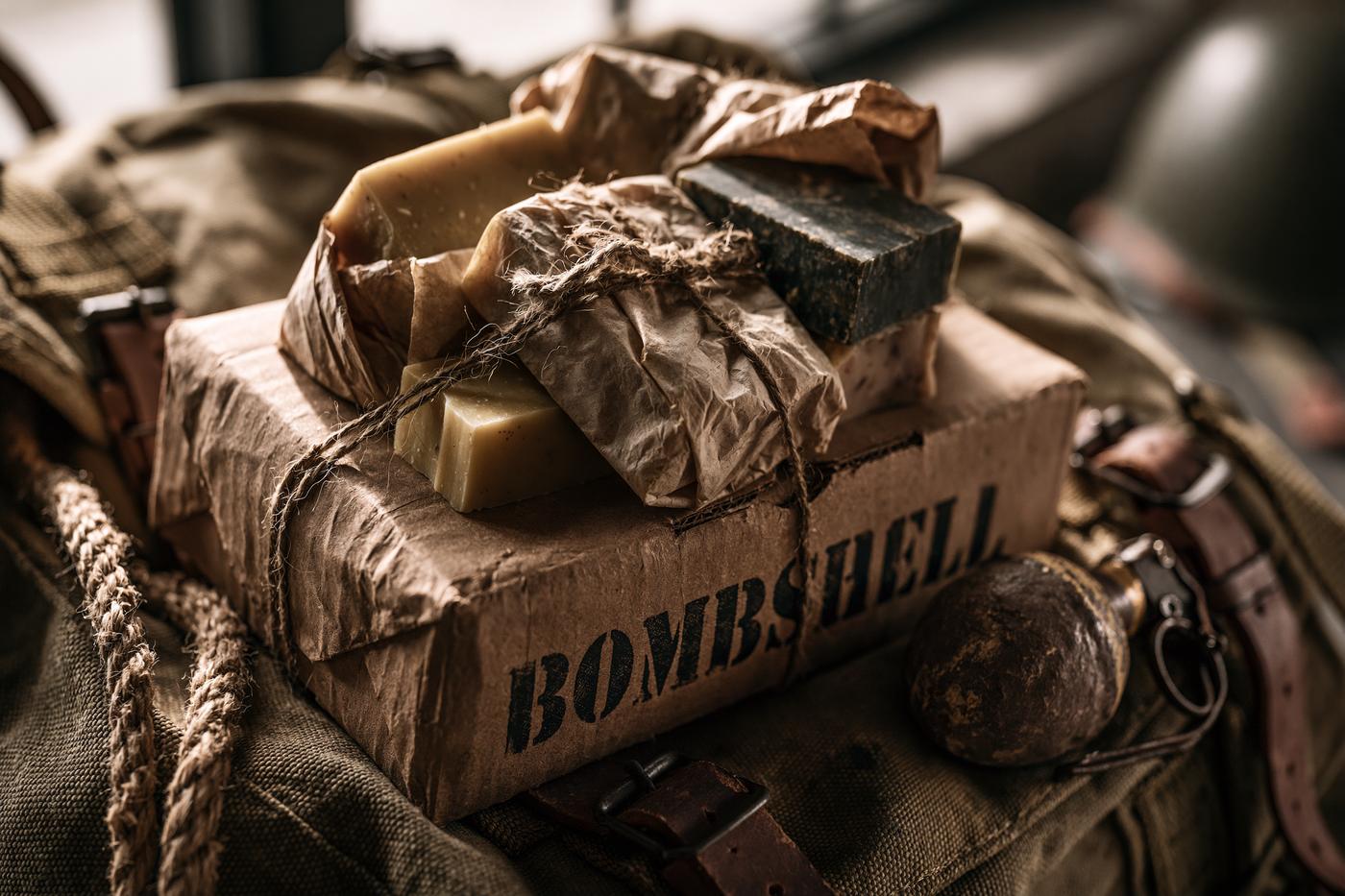 Bombshell Suds care package — soap bars and twine-tied kraft parcels stacked on a stenciled box atop an olive drab rucksack